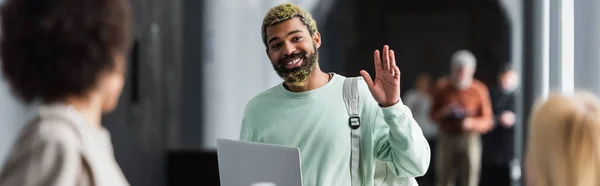 Smiling african american student with backpack and laptop waving at blurred friends in university, banner — Stock Photo