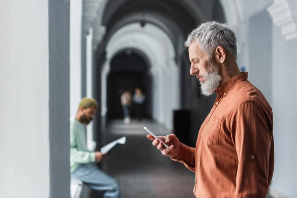 Side view of mature teacher using smartphone in corridor of university — Stock Photo