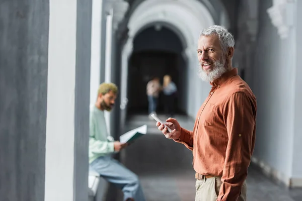 Professeur mature souriant utilisant un smartphone et regardant la caméra dans le couloir universitaire — Photo de stock