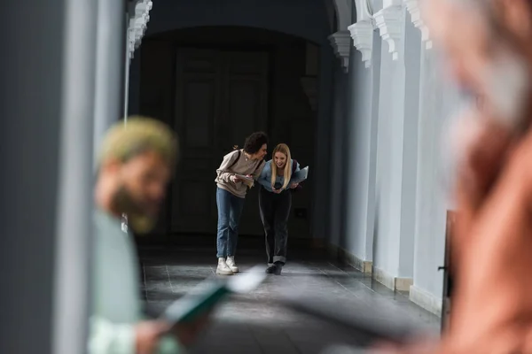 Estudantes multiétnicos alegres com notebook e laptop andando no corredor universitário — Fotografia de Stock