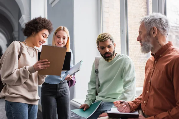 Positive Studenten mit Notizbüchern in der Nähe eines reifen Lehrers auf dem Flur der Universität — Stockfoto