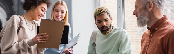 Sonriendo estudiantes interracial mirando cuaderno cerca de amigo y profesor en la universidad, pancarta - foto de stock