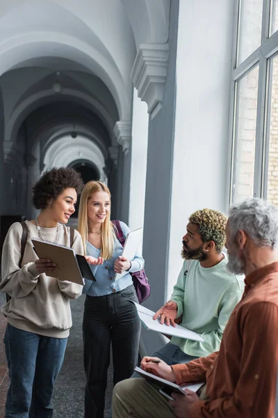 Позитивні багатоетнічні студенти тримають блокноти поруч з другом і вчителем в університетському коридорі — стокове фото