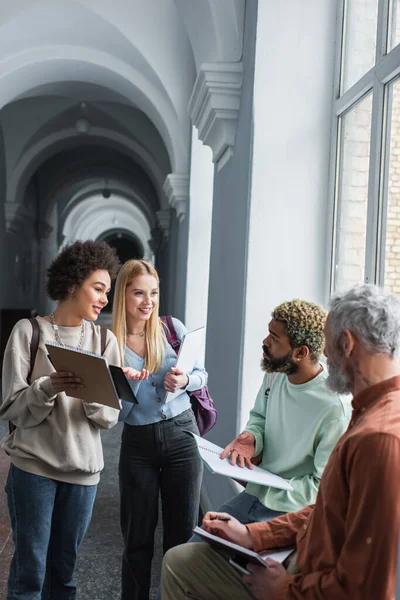 Positive multiethnische Studenten mit Notizbüchern in der Nähe von Freund und Lehrer im Universitätsflur — Stockfoto