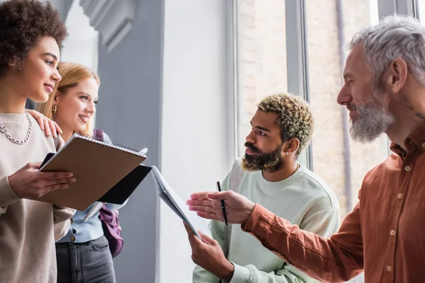 Mature teacher pointing with hand at notebook near smiling multiethnic students in university — Stock Photo