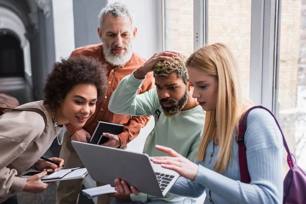 Interrassische Studenten schauen auf Laptop in der Nähe des Lehrers im Flur der Universität — Stockfoto