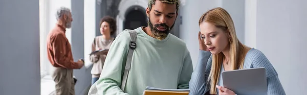 Afrikanischer Student hält Notizbücher in der Nähe von Freunden mit Laptop im Flur der Universität, Banner — Stockfoto