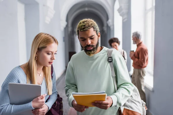 Estudantes inter-raciais segurando cadernos e laptop no corredor universitário — Fotografia de Stock