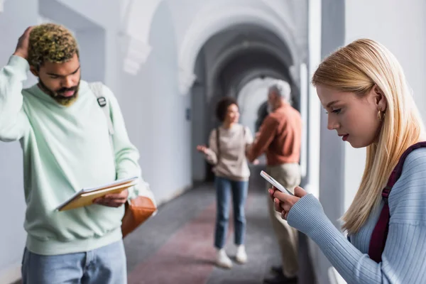 Studente utilizzando smartphone vicino sfocato amico afroamericano con quaderni nel corridoio universitario — Foto stock