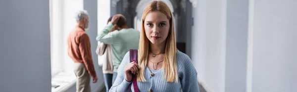 Blonde Studentin mit Rucksack blickt in die Kamera auf dem Flur der Universität, Banner — Stockfoto