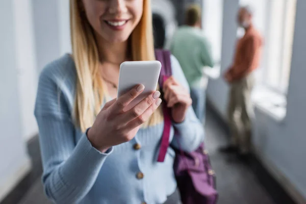 Cropped view of smiling student with backpack using smartphone in university corridor — Stock Photo