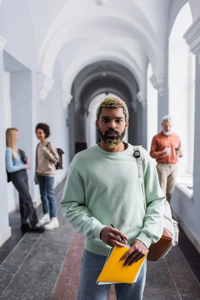 Studente afroamericano in possesso di quaderni e guardando la fotocamera nel corridoio dell'università — Foto stock