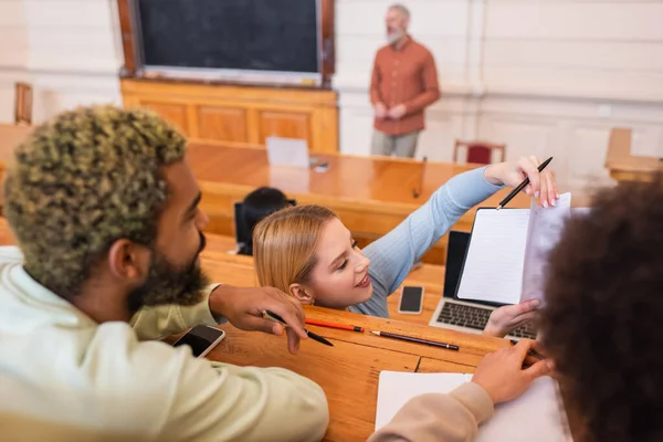 Smiling student holding notebook near blurred african american friends in university — Stock Photo