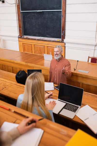 Professeur joyeux pointant avec la main à l'étudiant flou près des ordinateurs portables et des ordinateurs portables à l'université — Photo de stock