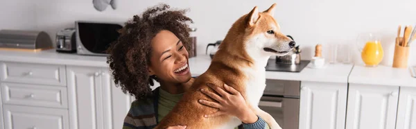 Веселая африканская американка, обнимающая шибу ину-собаку на кухне, баннер — стоковое фото
