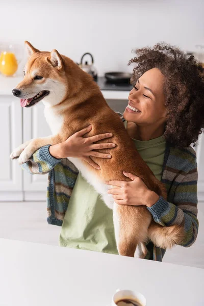 Alegre africano americano mujer holding shiba inu perro cerca café taza en cocina - foto de stock