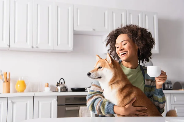 Laughing african american woman sitting in kitchen with cup of coffee and shiba inu dog — Stock Photo