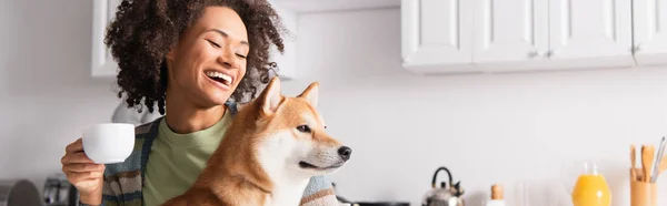 Riéndose africana americana mujer celebración mañana café cerca shiba inu perro, bandera - foto de stock