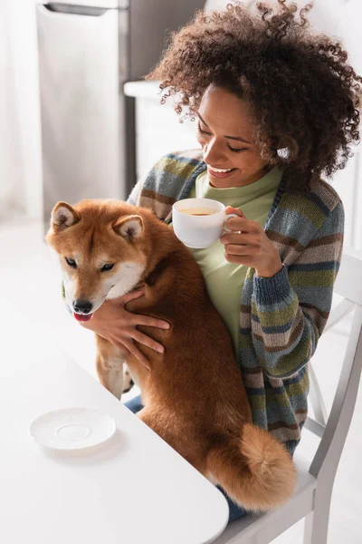 Sorridente donna africana americana che tiene in mano una tazza di caffè mentre coccola il cane shiba inu — Foto stock