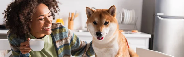 Shiba inu perro sobresaliendo lengua cerca de feliz africana americana mujer con taza de café, bandera - foto de stock