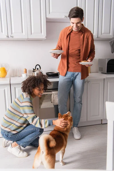 Smiling african woman stroking shiba inu dog near boyfriend with delicious pancakes — Stock Photo