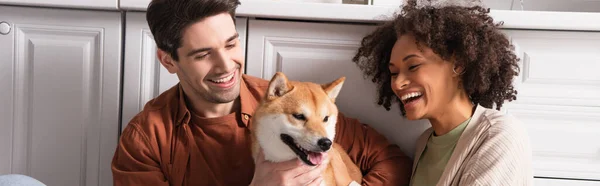 Felice coppia interrazziale sorridente vicino divertente shiba inu cane in cucina, banner — Foto stock