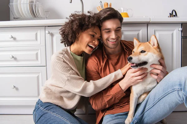 Pleased interracial couple hugging shiba inu dog while sitting on kitchen floor — Stock Photo