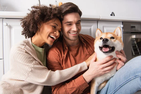 Cheerful man cuddling shiba inu dog near smiling african american girlfriend in kitchen — Stock Photo
