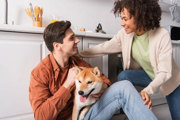 Feliz interracial pareja mirando uno al otro cerca divertido shiba inu perro en cocina - foto de stock