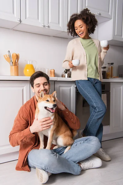 Homem alegre sentado no chão com shiba inu cão perto sorrindo afro-americano namorada com xícaras de café — Fotografia de Stock
