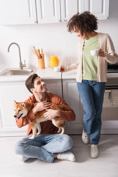 Cheerful african american woman holding morning coffee near boyfriend sitting on floor with shiba inu dog — Stock Photo