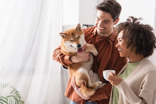 Allegro uomo in possesso di divertente shiba inu cane vicino afro americano donna con tazza di caffè — Foto stock