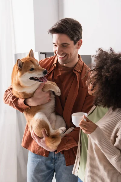 Homem alegre segurando shiba inu cão perto afro-americano namorada com xícara de café — Fotografia de Stock