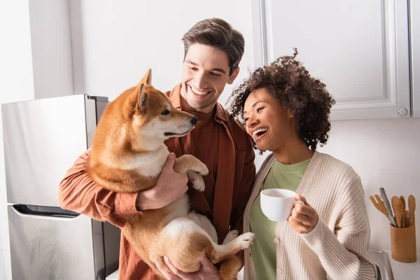 Lächelnder Mann kuschelt Shiba-Inu-Hund in der Nähe fröhlicher afrikanisch-amerikanischer Freundin mit Kaffeetasse — Stockfoto