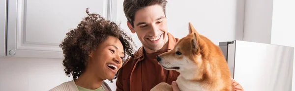 Engraçado shiba inu cão perto sorrindo casal interracial na cozinha, banner — Fotografia de Stock