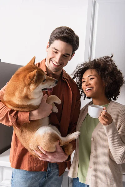 Smiling man holding shiba inu dog near happy african american girlfriend with morning coffee — Stock Photo