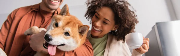 Borrosa pareja interracial sonriendo cerca shiba inu perro sobresaliendo lengua, bandera - foto de stock