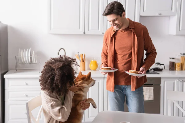 African american woman cuddling shiba inu dog near happy boyfriend with pancakes — Stock Photo