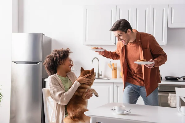 Africano americano mujer abrazo shiba inu perro cerca emocionado novio con desayuno - foto de stock