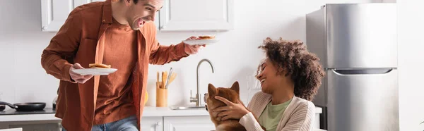 Homem surpreso segurando café da manhã perto de mulher ameican africana feliz com shiba inu cão, banner — Fotografia de Stock