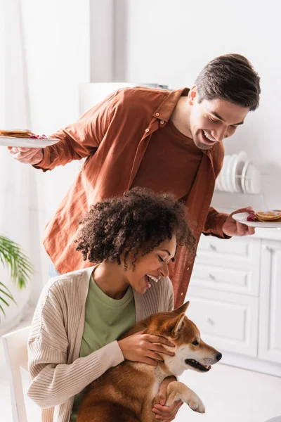 Espantado Africano americano mulher abraçando shiba inu cão perto sorrindo namorado com café da manhã — Fotografia de Stock