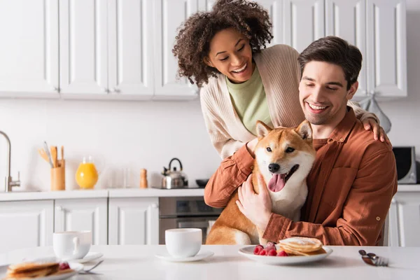 Uomo sorridente seduto con shiba inu cane vicino felice ragazza africana americana e gustosi pancake — Foto stock