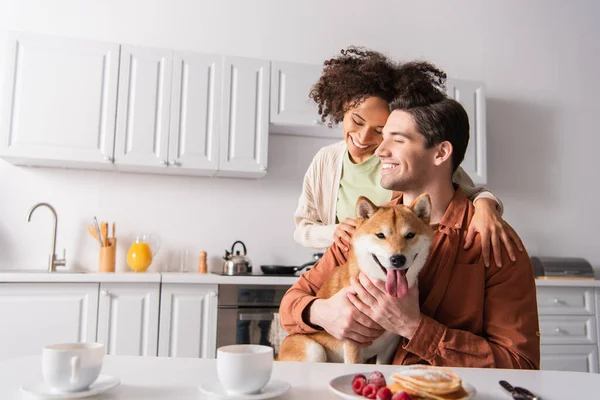 Felice coppia interrazziale sorridente con gli occhi chiusi vicino shiba inu cane e prima colazione in cucina — Foto stock