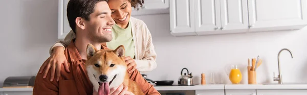 Shiba inu dog sticking out tongue near happy interracial couple in kitchen, banner — Stock Photo