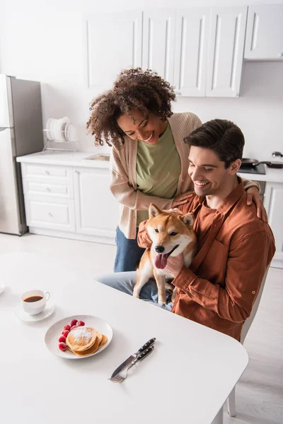 Glücklich Mann hält shiba inu Hund in der Nähe des Frühstücks und glücklich afrikanisch-amerikanische Freundin — Stockfoto