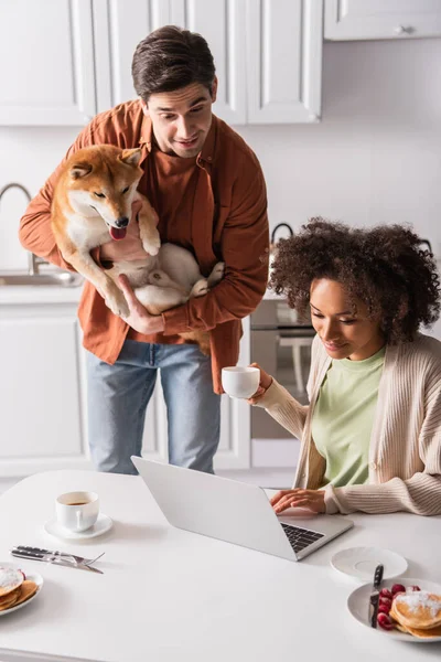 Homme surpris avec shiba inu chien regardant ordinateur portable près de petite amie afro-américaine avec tasse de café — Photo de stock