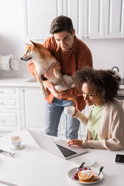 Young man with shiba inu dog looking at laptop near smiling african american girlfriend — Stock Photo