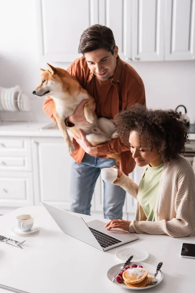 Jovem com shiba inu cão olhando para laptop perto sorrindo afro-americana namorada — Fotografia de Stock