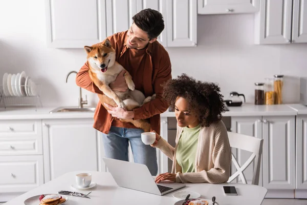 Happy man holding shiba inu dog near african american girlfriend working on laptop during breakfast — Stock Photo