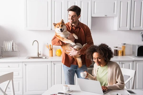 Homme souriant tenant shiba inu chien près afro-américaine copine travaillant sur ordinateur portable dans la cuisine — Photo de stock